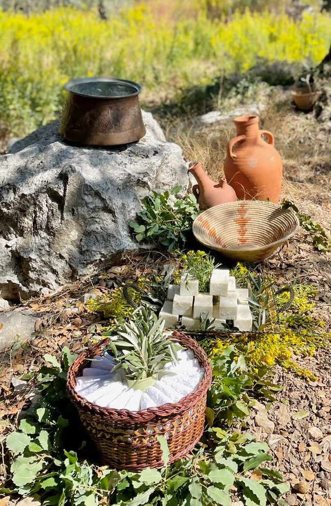 A scenic outdoor display featuring handmade soap, traditional clay jugs, a woven basket, and a copper pot on a large rock.