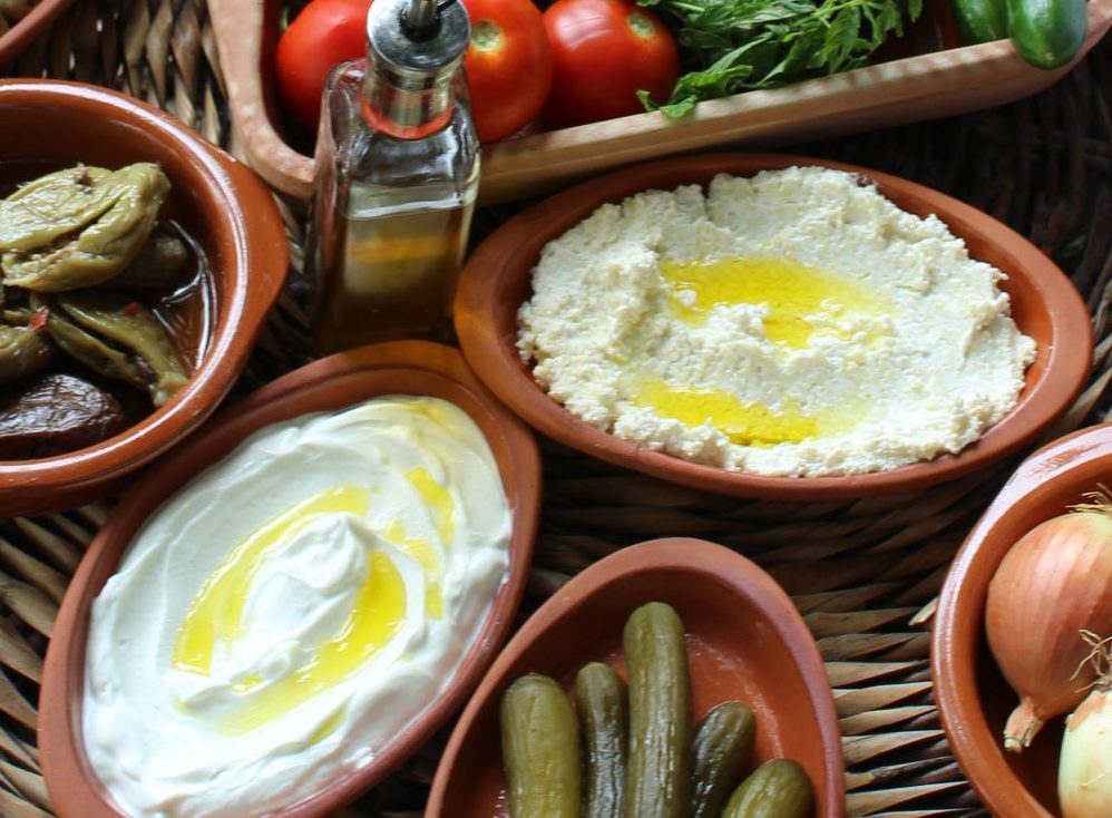 A close-up of traditional dishes including labneh with olive oil, pickles, and fresh vegetables arranged in clay bowls.